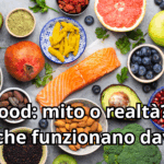 Composizione di vari superfood su sfondo scuro: trancio di salmone fresco, avocado, mirtilli, kiwi, broccoli, frutta a guscio, polveri di tè matcha e semi di chia. Al centro compare il titolo dell'articolo: Superfood: mito o realtà?