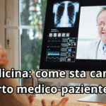 Vista sopra la spalla di un paziente che interagisce con un medico tramite videochiamata su un computer. Sul monitor sono visibili il volto sorridente del medico, una radiografia del torace e vari grafici di monitoraggio dei parametri vitali. Il titolo recita: 'Telemedicina: come sta cambiando il rapporto medico-paziente'