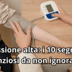 Inquadratura dall'alto di un braccio di una persona con indosso una camicia chiara, mentre un operatore sanitario posiziona il manicotto di un misuratore di pressione elettronico da tavolo. Sulla scrivania in legno si vedono anche un tablet e una penna. In primo piano compare il titolo: 'Pressione alta: i 10 segnali silenziosi da non ignorare'.
